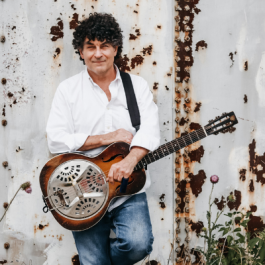 Chris Vallillo, standing in front of an aged wall holding a resonator guitar