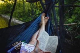 Person laying on a blue hammock in the woods reading a book