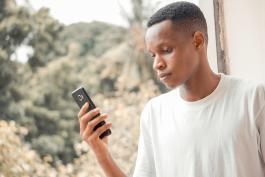 Photo of a black man in a white tee shirt holding a mobile phone with landscape in the background
